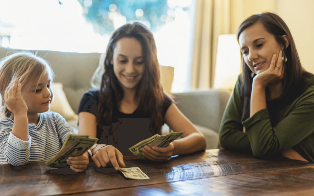 Two young girls sit at a table with their mother counting money. 