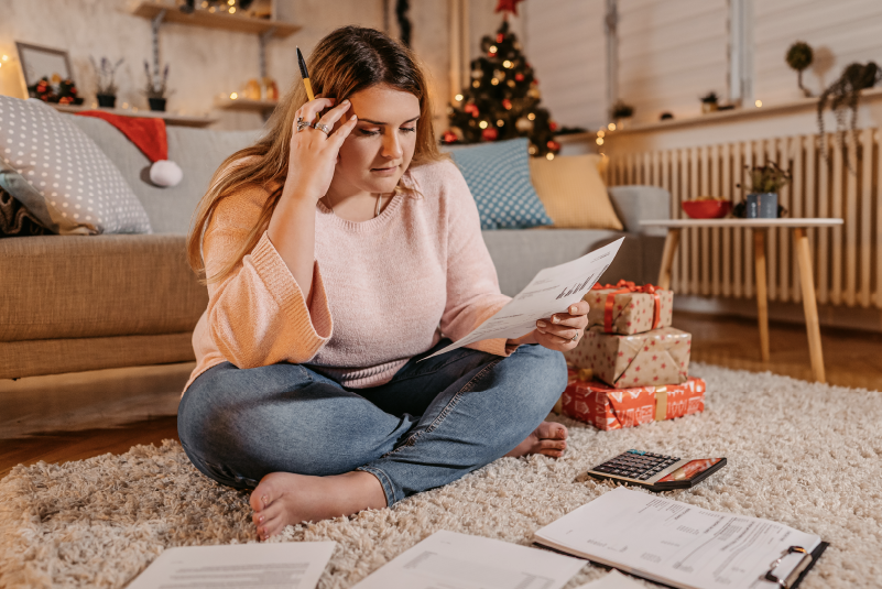 Woman sits crosslegged on the floor with financial papers spread out around her. Behind her are holiday gifts and a Christmas tree.