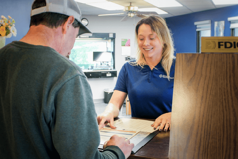 A man in a green sweatshirt and cap is signing a document at the counter while talking to a female banker in a blue shirt.