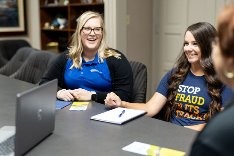 A group of FMB employees sit around a conference table with notepads and laptops. One wears a shirt that says, stop fraud in its tracks.