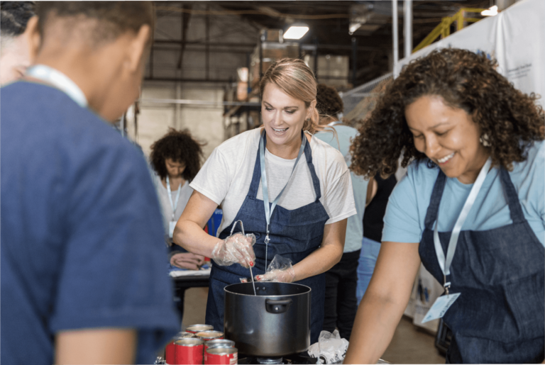 A woman wearing an apron stirs a pot at a soup kitchen while another woman dishes food to a young man.